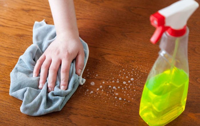 A close-up shot of a hand using a microfiber cloth and a spray bottle with cleaner to wipe down a hardwood floor finish.